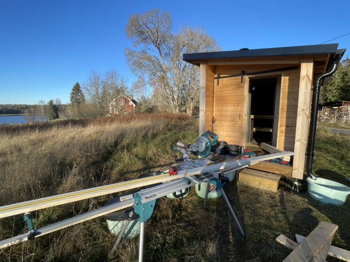 Nybyggd bastu av bondsågat virke på balkärra med tak och solpanel, vid sjö med verktyg framför.