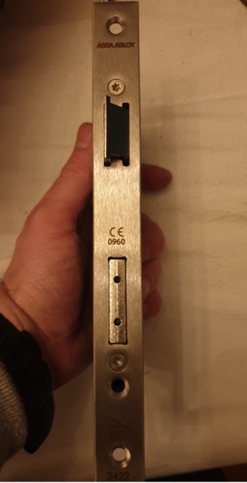Hand håller ett låshus från ASSA ABLOY med etiketten "CE 0960" och en synlig spärrbult utan nålshål.