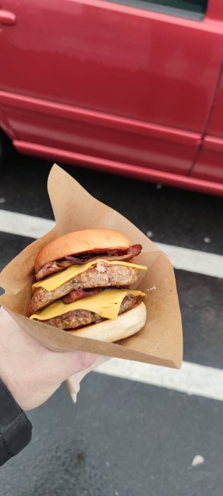 En hand som håller en hamburgare med två köttbullar, ost och bacon framför en röd bil.