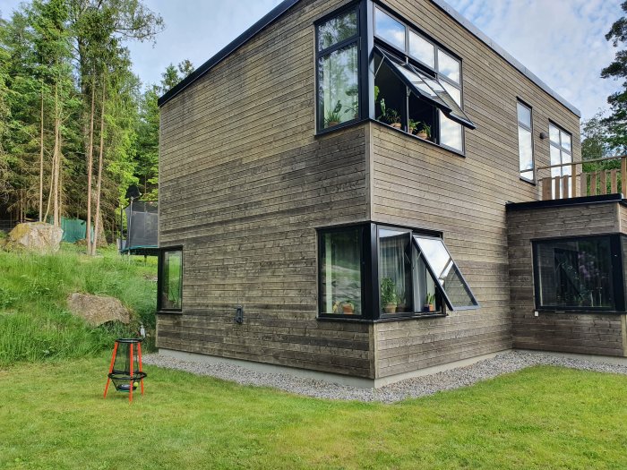 Baksidan av ett modernt hus med mörk träpanel och stora fönster, omgivet av grönska och en gräsmatta.