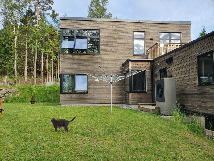Modern tvåvånings trävilla med balkong och stora fönster, omgiven av en grön gräsmatta och en katt i förgrunden.