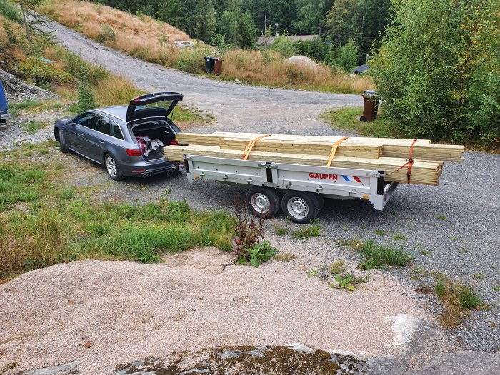 Bil och släpvagn lastade med trävirke för balkongbygge parkerade vid en grusväg.