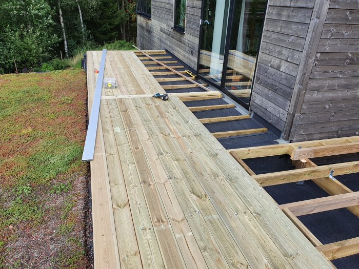 Pågående bygge av en balkong med ny lagd trägolv, kilade fästen, utan räcke, bredvid en sedumtäckt tak.