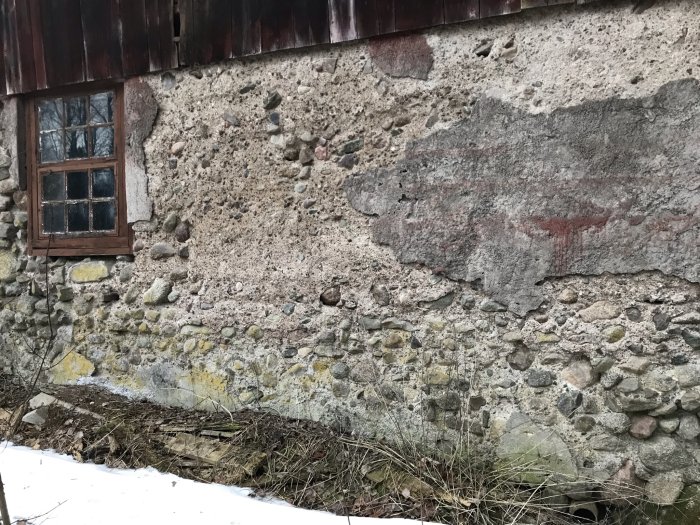 Sliten ladugårdsvägg av sten med partiellt avfallen puts, olika stenstorlekar, med fönster och snö i förgrunden.