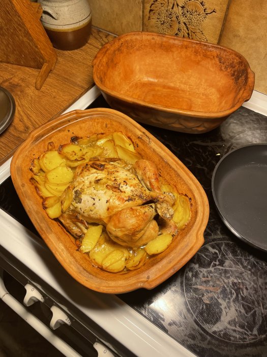 Tillagad kyckling med potatis i en öppen terrakotta-lergryta på köksbänk.