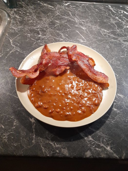 En tallrik med stekt bacon och bönor i tomatsås på ett marmorbord.