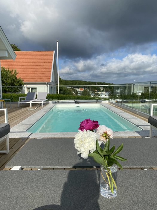 Uteplats med swimmingpool och solstolar, bukett pioner i förgrunden och mörka moln i himlen.