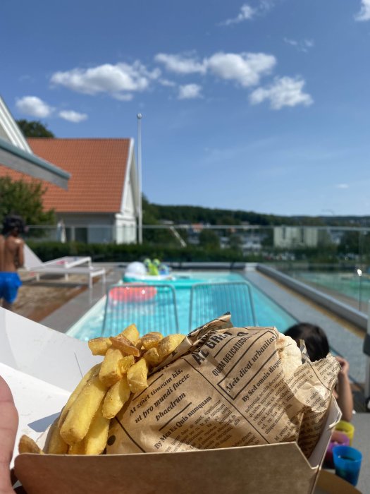 Kartong med pommes frites framför en blurrig pool och hus med blå himmel och solsken i bakgrunden.