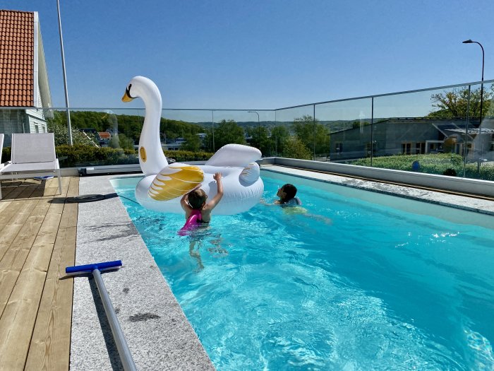 Utepool med klarblått vatten omgiven av ett glasstaket och trätrall, med en stor svanflotte och två personer som simmar.