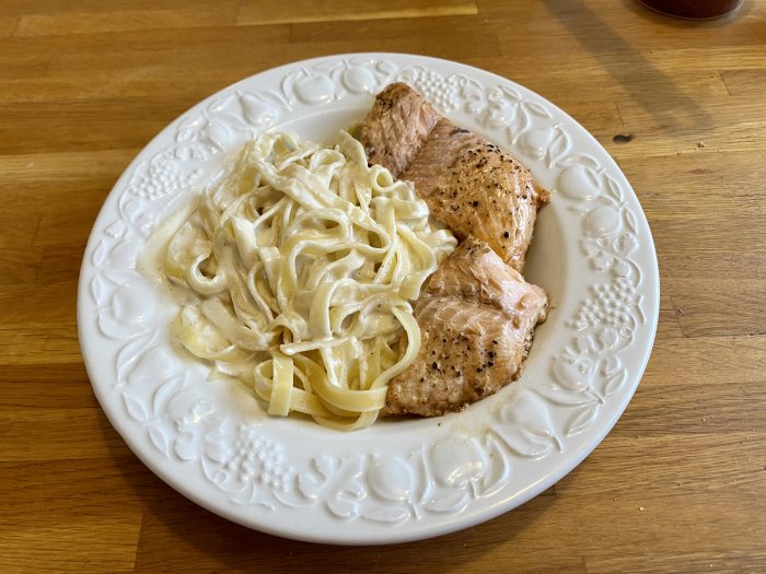 Tallrik med pasta i vitvinssås och stekt lax på ett träbord.