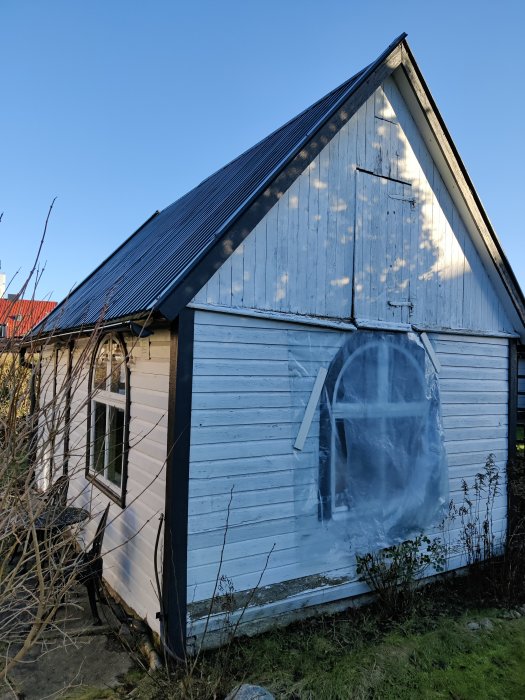 Liten blått och vitt målad stuga med sadeltak och plastöverdraget fönster i dagsljus.