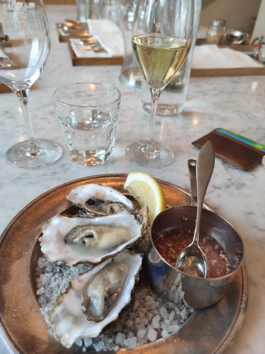 Ostron på is med citron och sås, ett glas vin och vattenglas i bakgrunden.