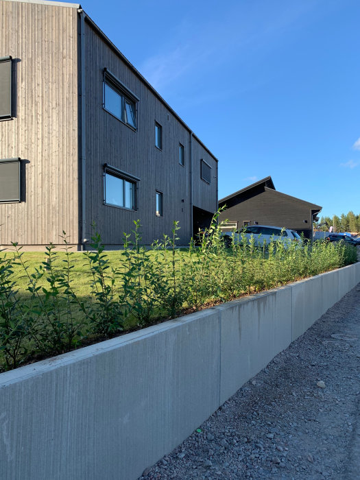 Modernt grått hus med svart tak och integrerad carport, omgivet av en nyplanterad häck och en nyligen konstruerad stödmur i förgrunden.