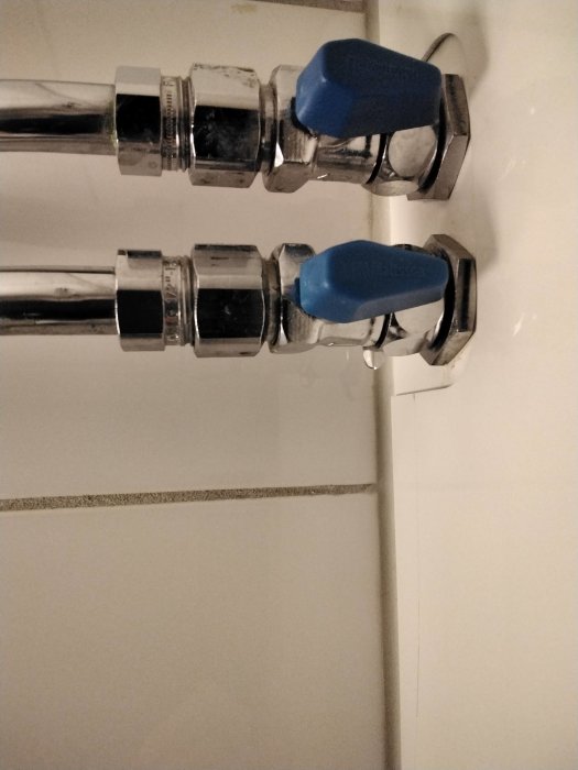 Närbild av två kromade varm- och kallvattenventiler med blå handtag monterade på vit kakelvägg.