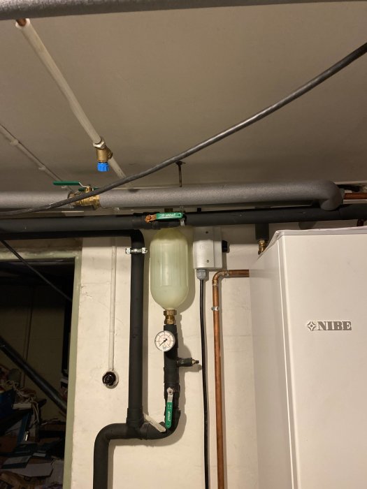 Pelletspanna med kopplade rör och en expansionskärl, tryckmätare och flera ventiler nära en NIBE värmepumps avstängd enhet.