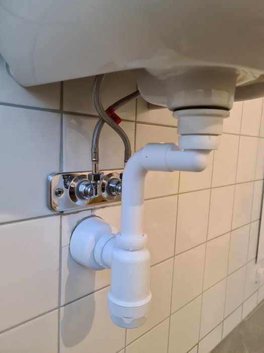 Nyinstallerad vattenlås och rörsystem under handfat med kakelvägg i bakgrunden.