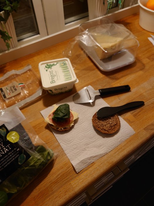 En smörgås under tillagning på ett köksbord med ost, pålägg och spenatblad, bredvid smör och knivar.