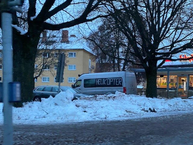 Vit skåpbil med texten "HELIKOPTER" parkerad vid snötäckt vägkant med träd och byggnader i bakgrunden.