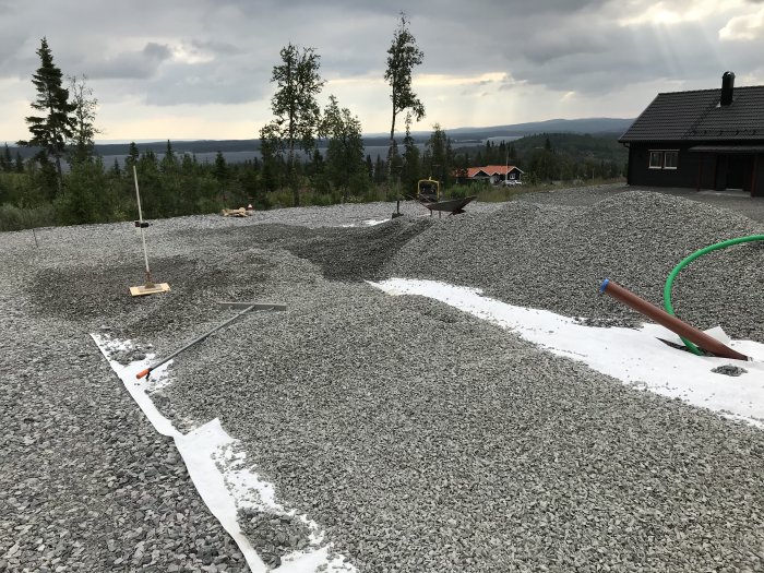 Grusbelagd byggarbetsplats med markduk, verktyg för anläggningsarbete och omgivande natur i bakgrunden.