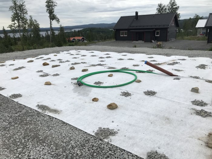 Markduk med utspridda stenar och en grönslingad slang på en byggplats, med hus i bakgrunden.