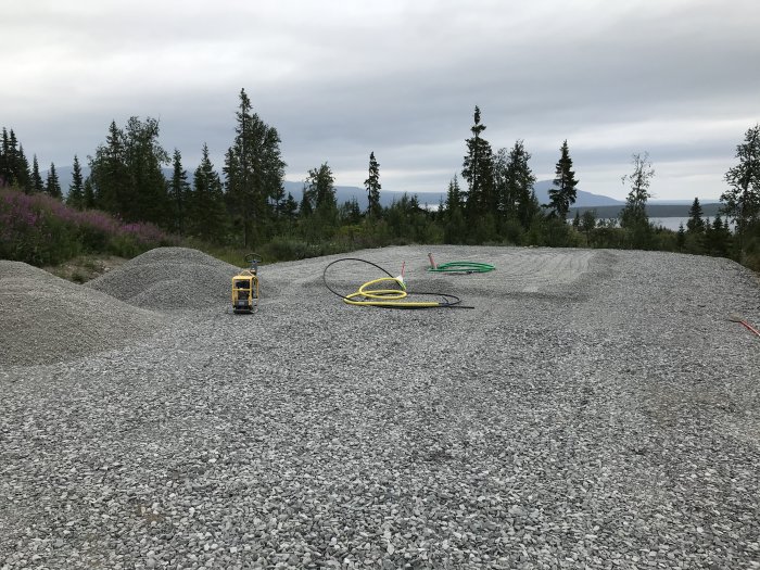 En byggplats med grus och markduk, utrustad med en laserinstrument och färgglada mätslangar, omgiven av skog och med en sjö i bakgrunden.