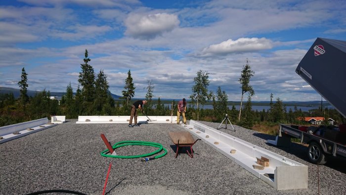 Två personer förbereder grund med grus och kantelement för byggprojekt med naturskön bakgrund.