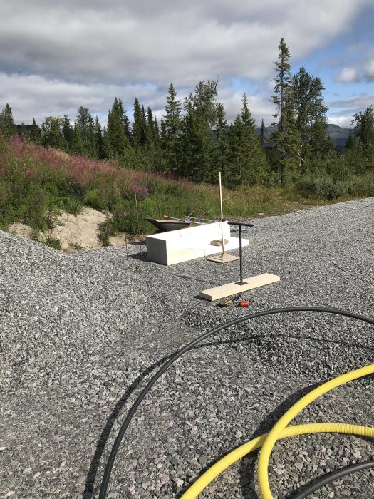 Byggplats med grusbädd, verktyg och kantelement förberedda för grundläggning, omgiven av natur.