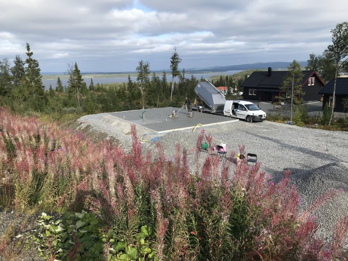 Byggplats med nylagt bärlager och grusbil som tippar material, omgivet av blommande växtlighet.