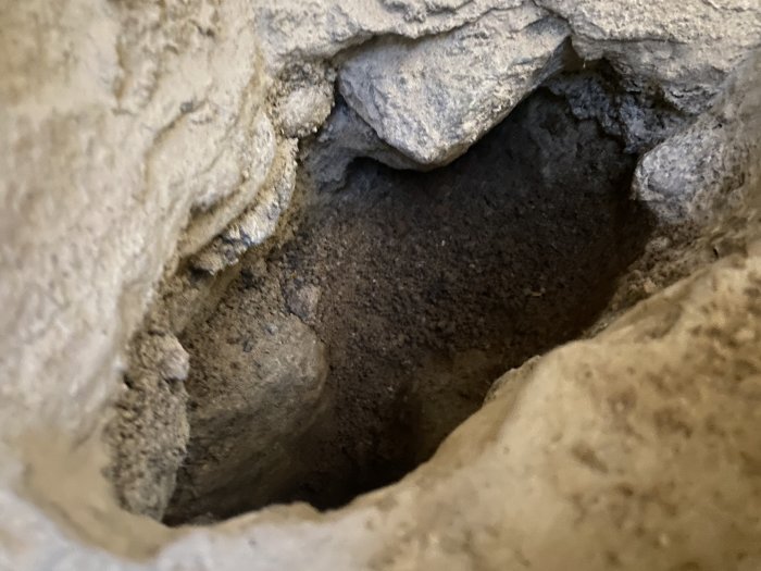 Uppbrutet betonggolv i ett hörn som avslöjar ett underliggande lager av större stenar och sand, troligtvis ett bärlager.