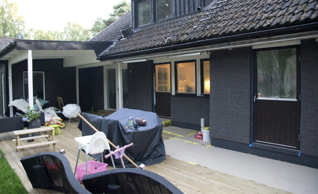 Ett hus med svart tegel och vita fönster efter renovering, med en nybyggd trädäck och barnleksaker.