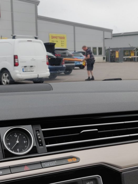 Bil lastar långt föremål vid Byggmax, man i förgrunden, osäker lastsäkring.