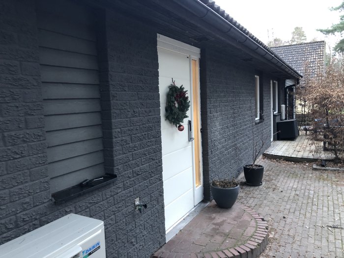 En husfasad med mörkgrå färg och en vit dörr, något dammig men inte blekt, med en julkrans på dörren.