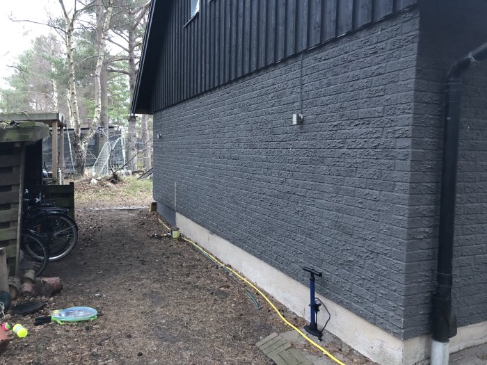 Mörkgrå fasad på hus med tecken på damm och pollendepositioner, omringat av lösa föremål och natur.