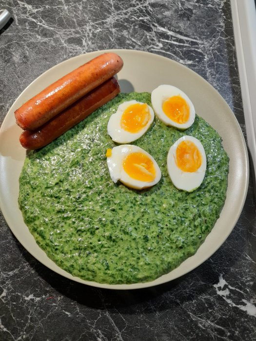 En tallrik med stuvad spenat, två halvor av ett kokt ägg och en grillad korv på en marmorbänk.