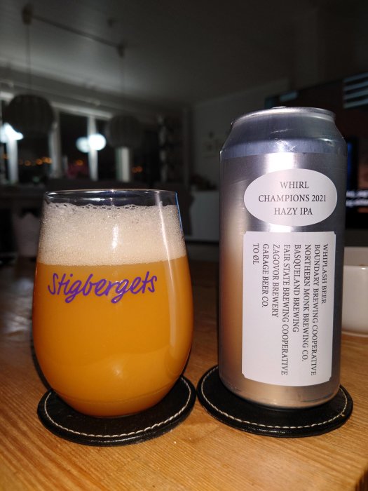 En ölburk märkt "WHIRL CHAMPIONS 2021 HAZY IPA" bredvid ett ölglas fyllt med skummande öl på ett bord.