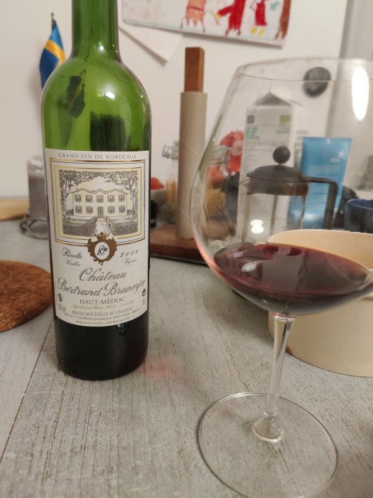 Flaska av Grand Vin de Bordeaux på ett bord bredvid ett vinglas med rödvin, inomhusmiljö.