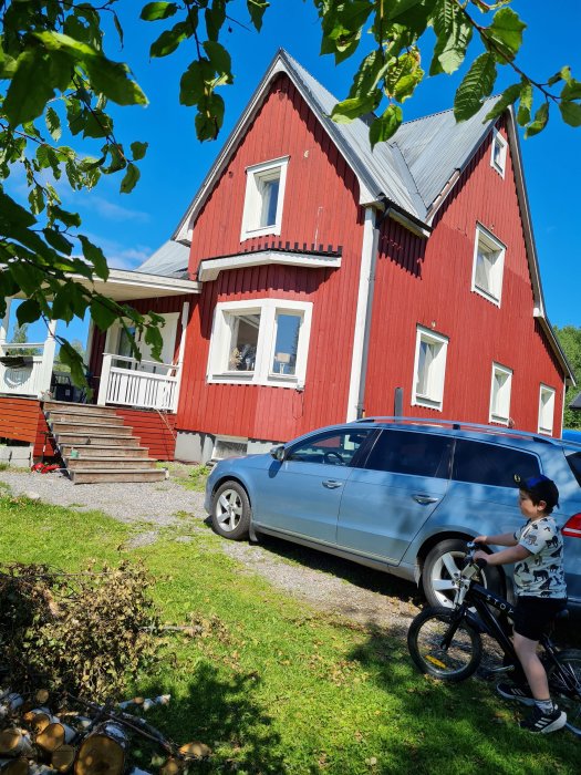 Rött trähus med vita fönster och veranda, en bil parkerad vid sidan och ett barn på cykel framför.