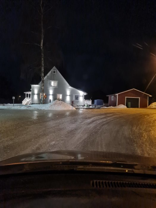 Utsikt från bil över snöig uppfart mot upplyst hus och förrådsbyggnad på kvällen.