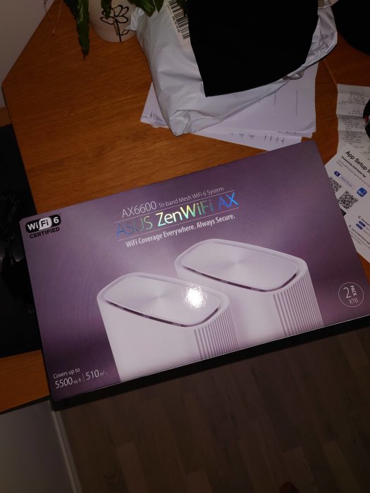 Förpackning för ASUS ZenWiFi AX, tri-band mesh Wi-Fi 6 system, på ett bord med oidentifierbara papper runtomkring.
