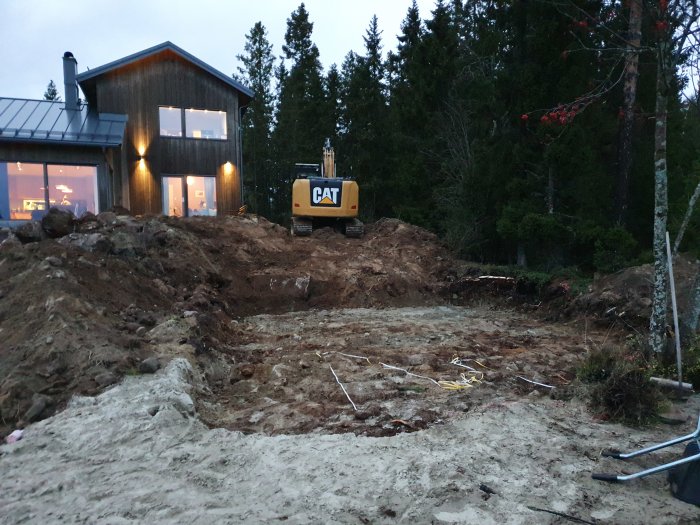 Grävmaskin vid en tom tomt med uppgrävd jord och ett hus i bakgrunden vid skymningen.