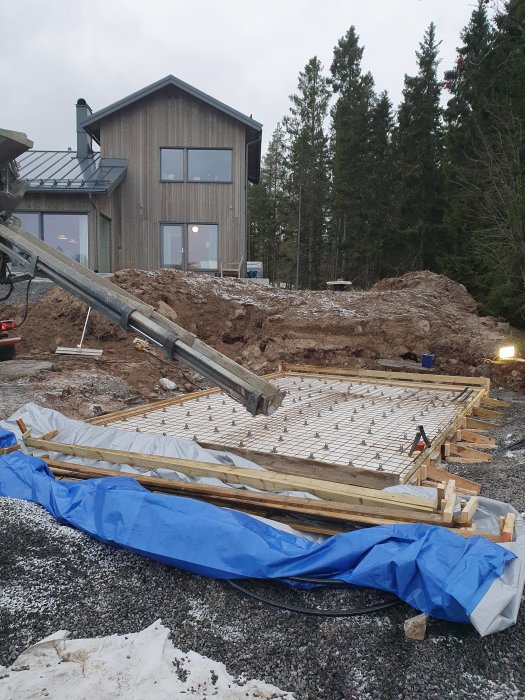 Grundläggning för utekök med armeringsjärn på träformer vid ett hus med skog i bakgrunden.