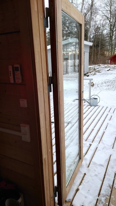 Utsikt genom öppen trädörr mot en snötäckt altan och trädgård.