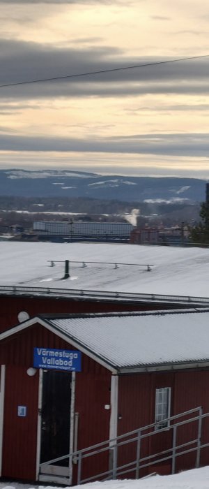 Röd värmetuga med snö på taket, skylt "Värmestuga Vallbacksbod", utsikt över snötäckt landskap och molnigt himmel.