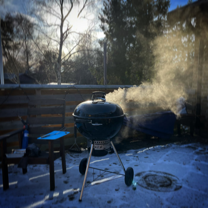 Grill på en snöig uteplats avger rök i solljus med träd i bakgrunden.