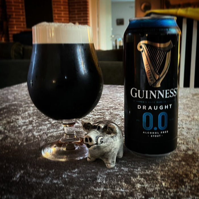 Ett glas mörkt alkoholfritt öl bredvid en Guinness 0.0 burk och en liten grisfigur på bordet.