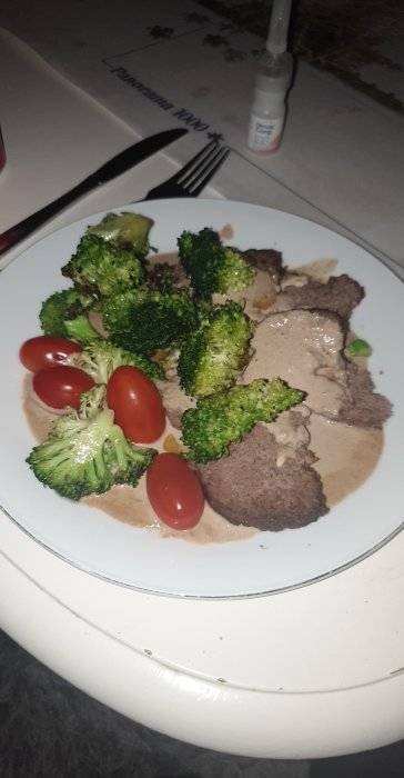 Köttfärslimpa på älgkött med gräddsås, omgiven av broccoli och körsbärstomater på en vit tallrik.