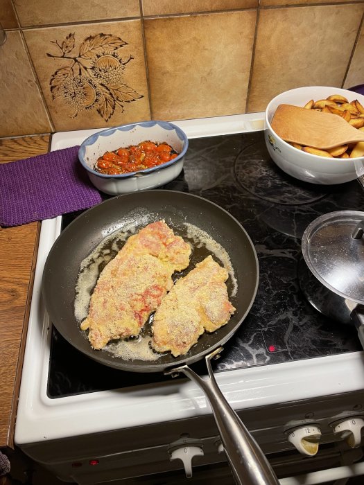 Panerade köttstycken steks i en stekpanna på spisen med tillbehör i närheten.