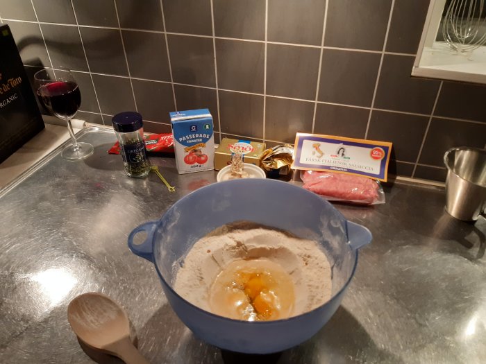 Ingredienser för pastatillagning med mjöl, ägg i skål, kött, tomatsås och kryddor på köksbänk.