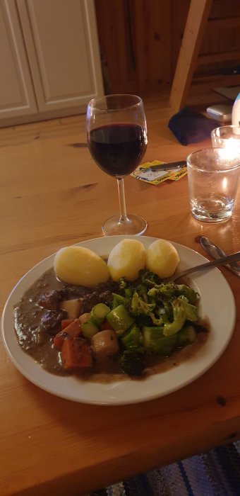 Tallrik med hjortgryta, broccoli och potatis bredvid ett glas rött vin och ett glas vatten.