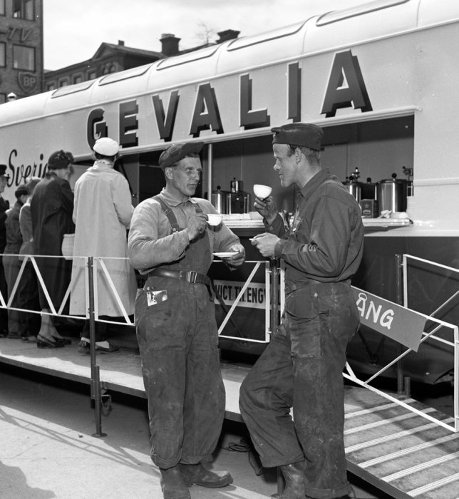 Svartvit bild av 1950-tals Gevalia kaffeservering med män som dricker kaffe och en lastbil med texten "GEVALIA".